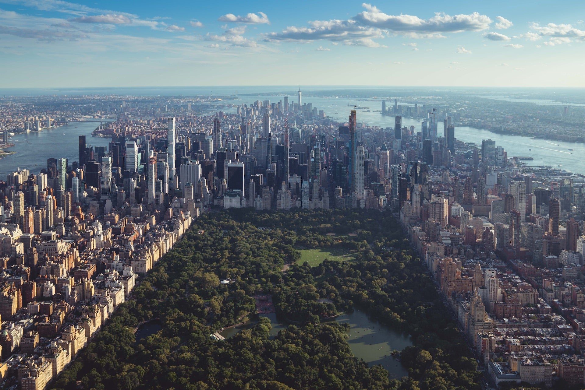 An aerial view of Central Park — one of the best parks for e-bikes — with lots of green grass and trees, surrounded by the rest of Manhattan on a sunny day with blue skies and white clouds