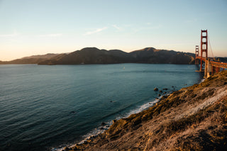 A warm sunny view of the Golden Gate Bridge over San Francisco Bay, featuring rocky hills and shoreline with mountains in the distanc
