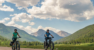 Two riders on Magnum fat tire e-bikes look at the scenery surrounding them: rolling green hills, mountains in the background, and a blue sky with fluffy white clouds.