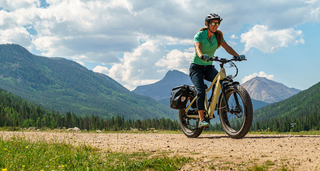 A rider in cyan polo and dark blue cropped pants rides a beige Magnum Scout e-bike on a flat dirt path among lush green mountains