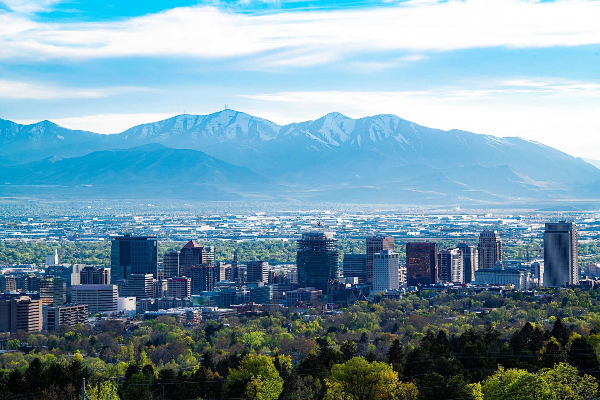 A high view of Salt Lake City, Utah on a bright sunny day with trees in the foreground, mountains in the background, and the city in the center of the frame