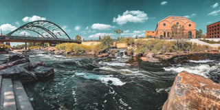 The iconic natural landscape of Denver Colorado featuring a rushing river alongside a bank with trees, a bridge crossing the river, and a brick building