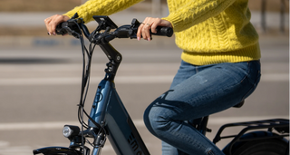 A woman in a yellow sweater and dark denim jeans grips the handlebars of a blue Cosmo X e-bike with her hands on the brakes. The road is out of focus in the background.