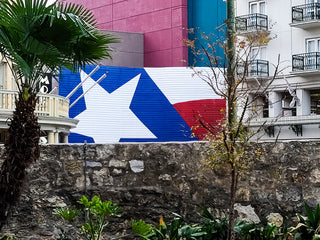  Behind a stone wall is a bright mural of the Texas state flag nestled between white buildings in the Alamo plaza