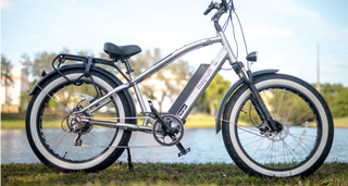 A silver Magnum Ranger e-bike stands parked by a river on a sunny day; due to the angle, light is visible between the front tire and fender on the bike