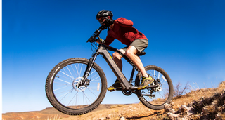 Rider in red long-sleeve shirt and khaki shorts with black sunglasses and black bike helmet rides a black Magnum Summit e-bike over a small jump on rocky terrain