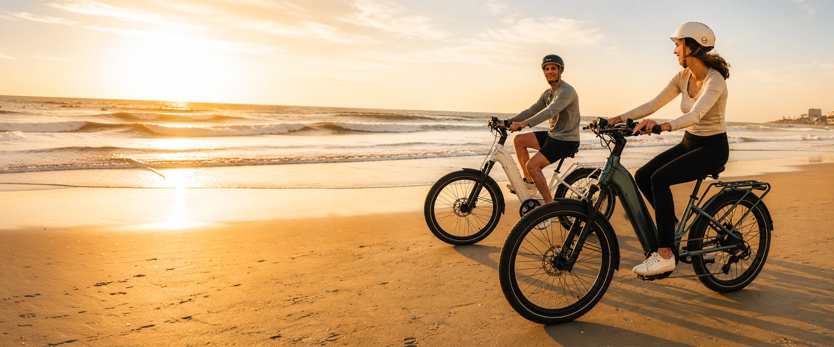 Two people riding electric bicycles on a sandy beach during a golden hour sunset, with ocean waves and the sky in the background.